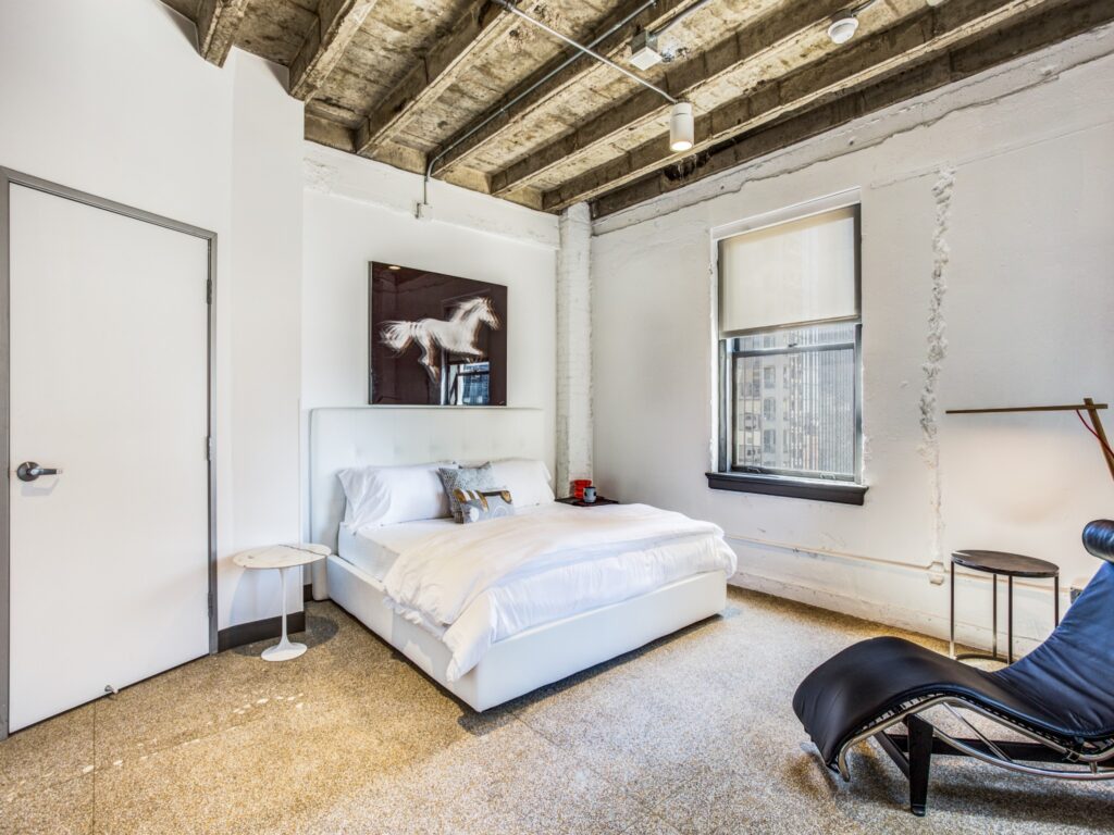 Bedroom with hard flooring, exposed wooden rafters above bed, and two windows with view of downtown Dallas.