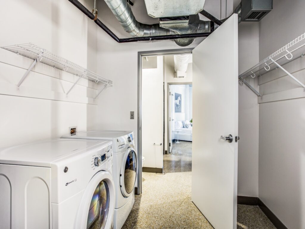 Apartment laundry room with washer and dryer, exposed industrial venting above, and shelving system on both walls.