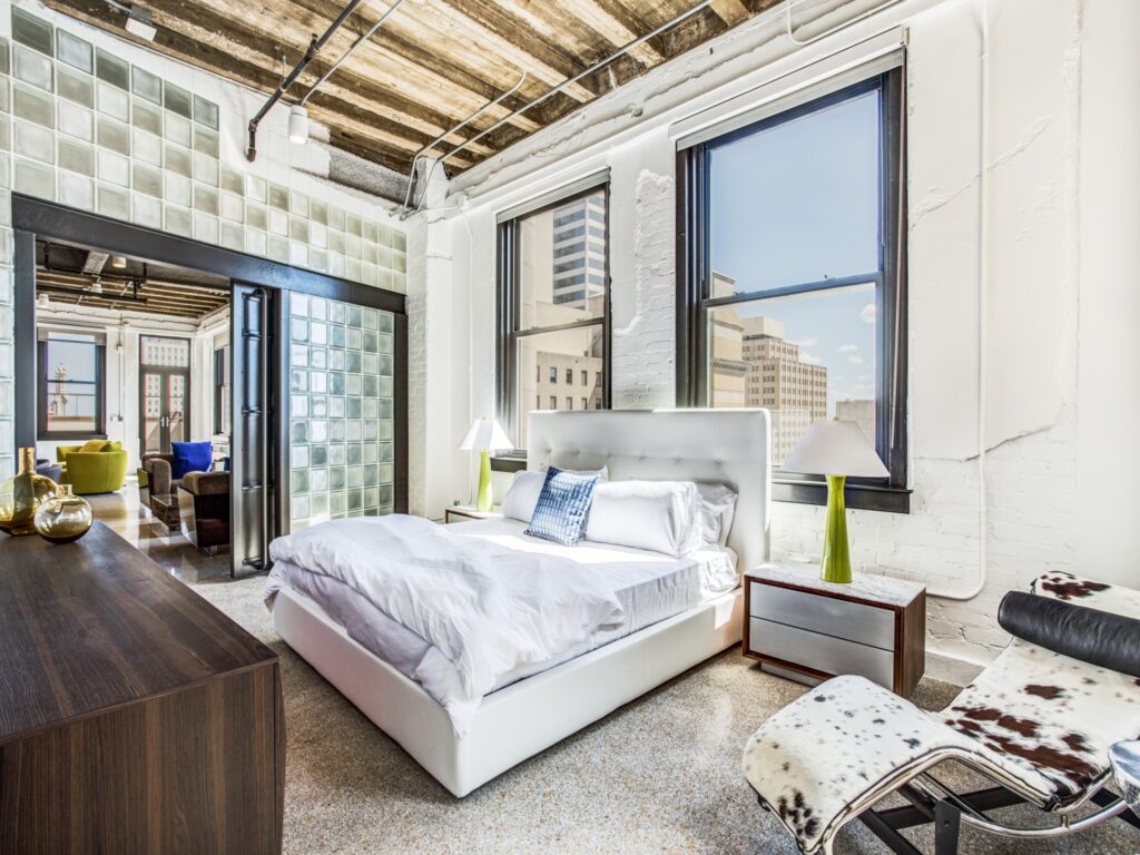 Bedroom with hard flooring, exposed wooden rafters above bed, decorative glass wall with sliding barn door, two windows with view of downtown Dallas.