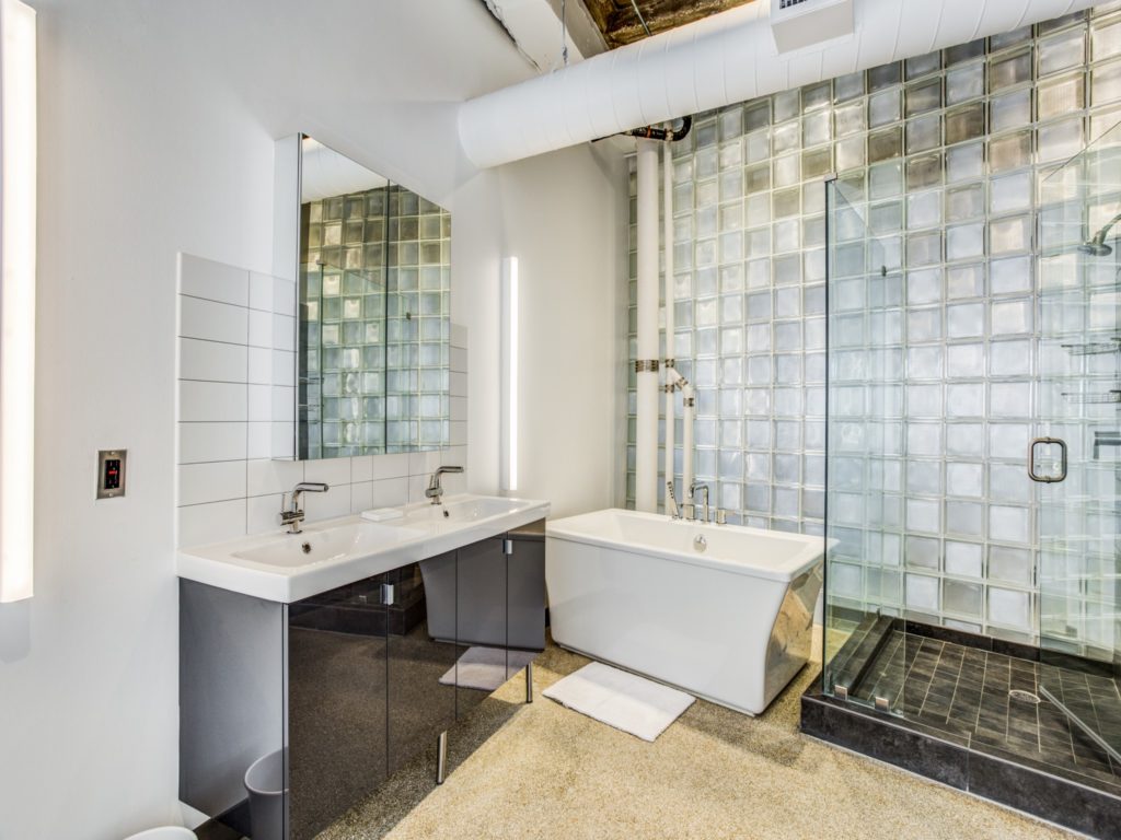 Bathroom with double vanity sink area, stand-up glass shower with ceramic tiling, large rectangle shaped soaking tub, exposed venting and pipes in corner.